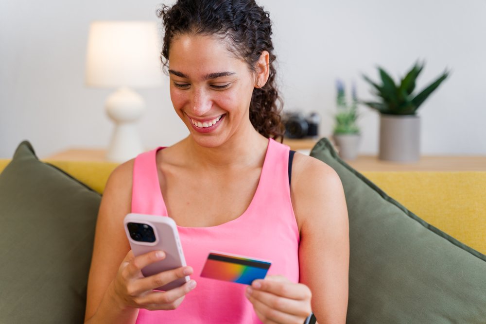 A woman in a pink sleeveless top happily holds a smartphone and a credit card while sitting on a couch at home, Spain.