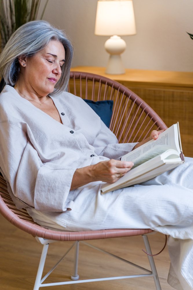 Middle-aged woman with gray hair reads a book while seated comfortably indoors in Spain under warm light.