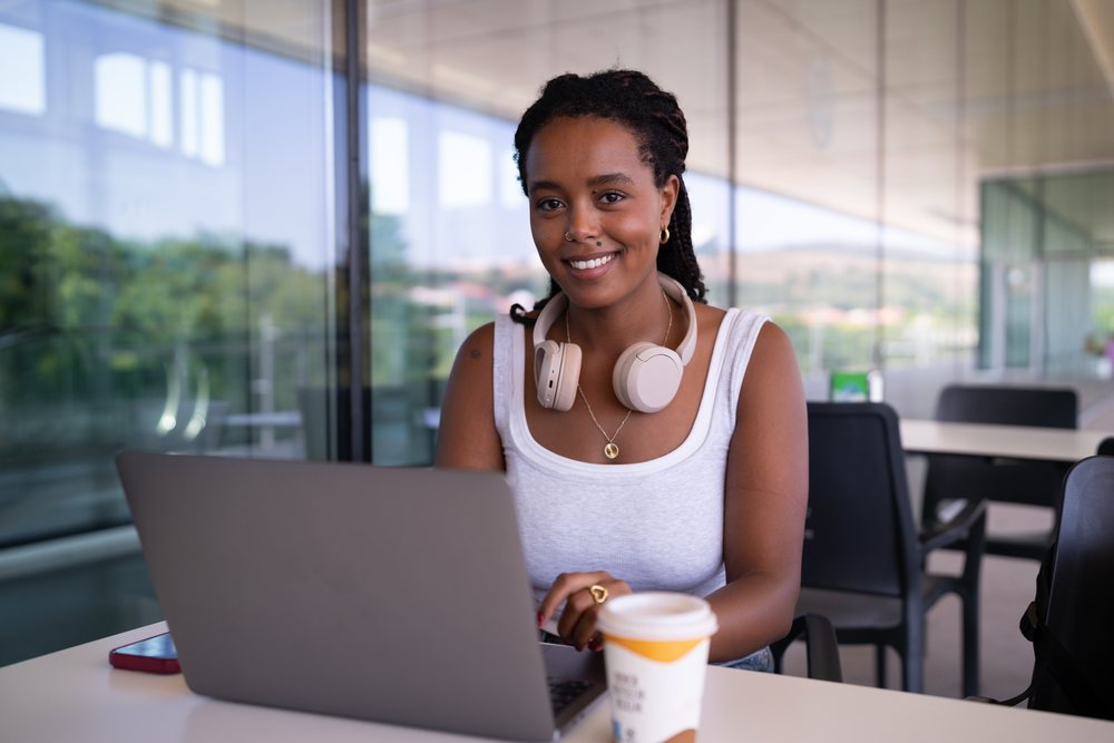 A young black woman wearing headphones works on a laptop with a coffee cup nearby in a modern office, remote work, travel, Santiago De Compostela, Spain.