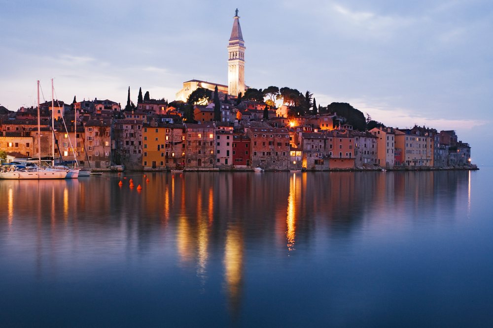 The town of Rovinj's illuminated waterfront and church tower reflect in calm water, Sunset, Croatia.