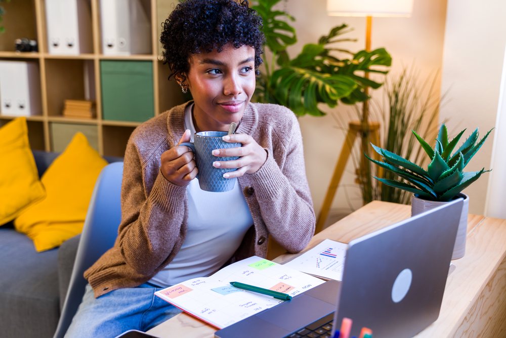 Woman sitting at a wooden desk holding a grey patterned coffee mug with laptop and calendar planner nearby in a cozy home office, Spain.