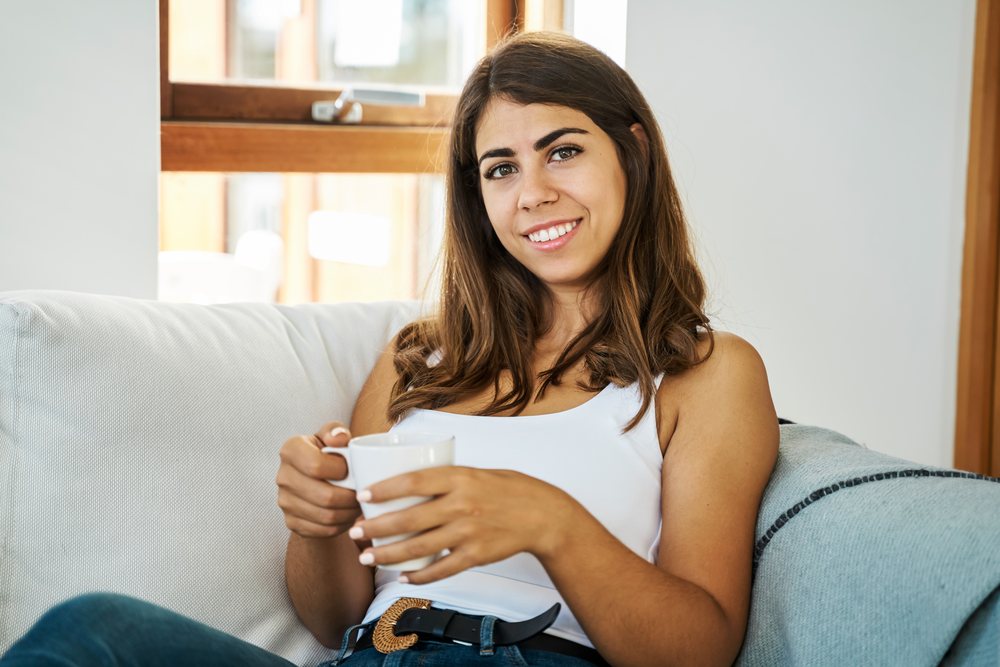 A white woman sitting comfortably indoors holding a cup and smiling