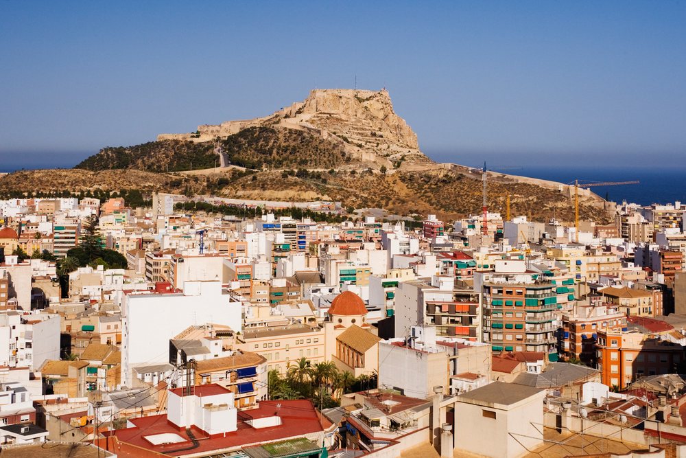 View of Castillo de Santa Barbara fortress atop a hill overlooking Alicante's urban residential buildings and the Mediterranean Sea under a clear blue sky, Valencia, Spain.