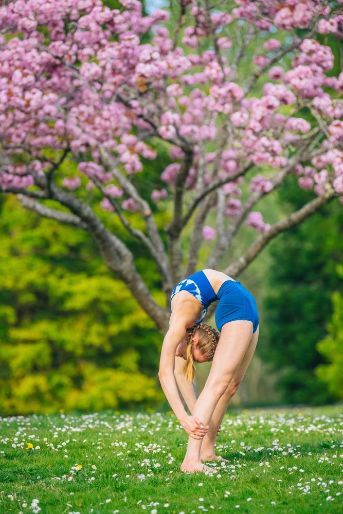 Woman in blue athletic clothing stretches barefoot outdoors on grassy ground with scattered daisies near a large pink blossoming tree in spring, USA.