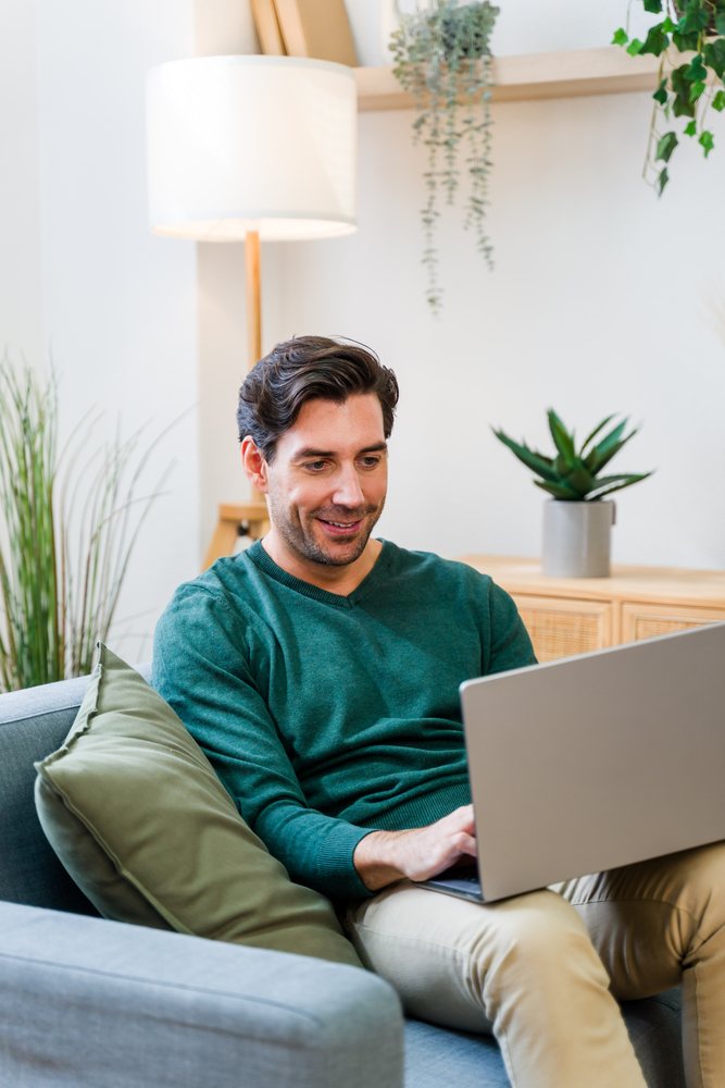 Man wearing green sweater and beige pants sitting on a sofa using a laptop in a modern living room with plants and a floor lamp, Spain.