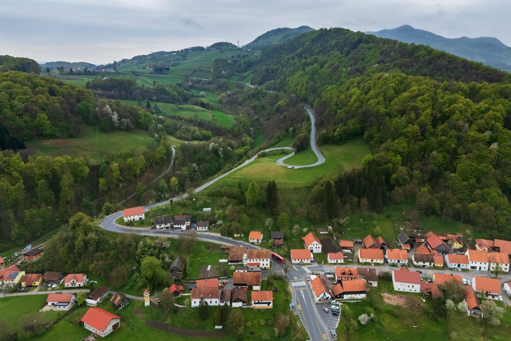 Curved winding road passes through the village of Podsreda with houses, red roofs, and green wooded hills in Slovenia's Kozjanski Regional Park, landscape, cityscape, Podsreda Market Town.