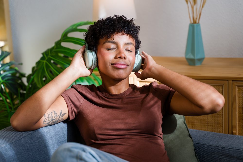 Woman with short afro curls wearing headphones and a brown shirt relaxes on a couch in a living room with plants and a vase nearby, Spain.