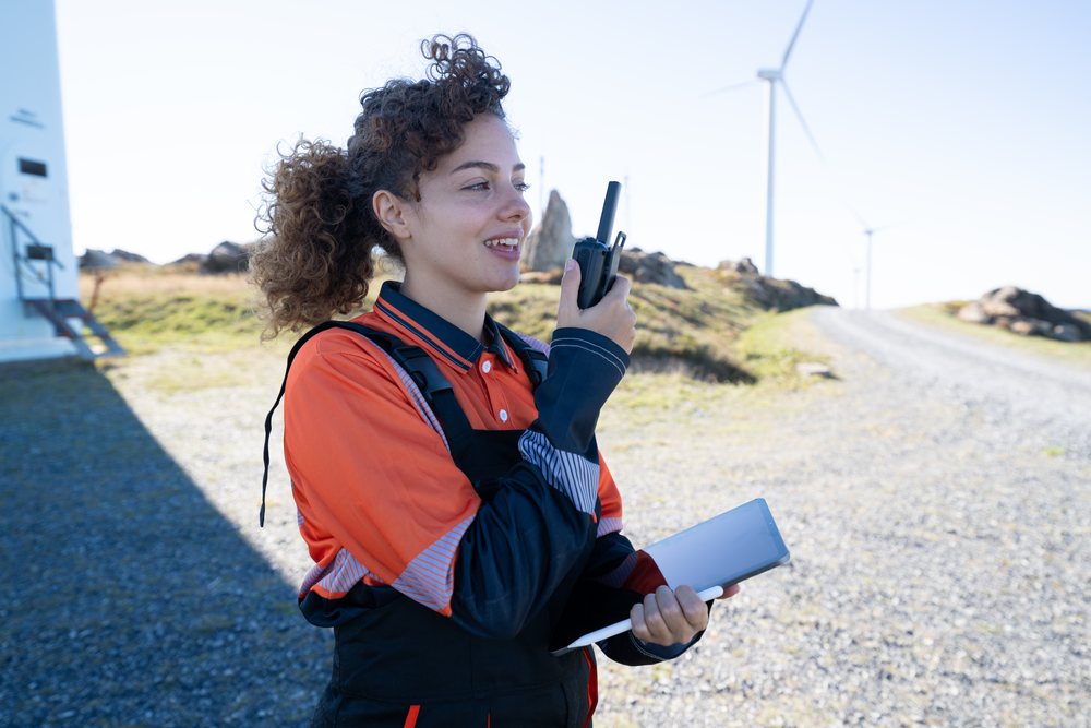 Woman in workwear a tablet and walkie talkie at a sunlit wind turbine site, engaged in renewable energy work, Galicia, Spain.