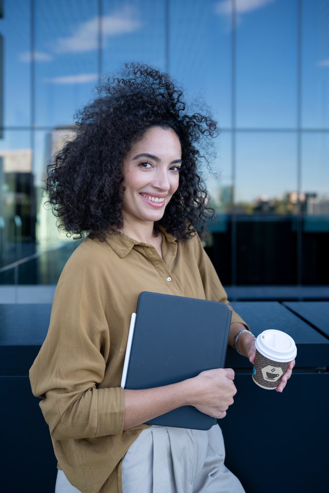 Smiling woman with curly hair holding a tablet and takeaway coffee outside an urban glass building, Madrid, Spain.