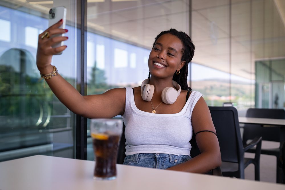 A woman wearing headphones takes a selfie with her smartphone while sitting at an outdoor table, remote work, travel, Santiago De Compostela, Spain.