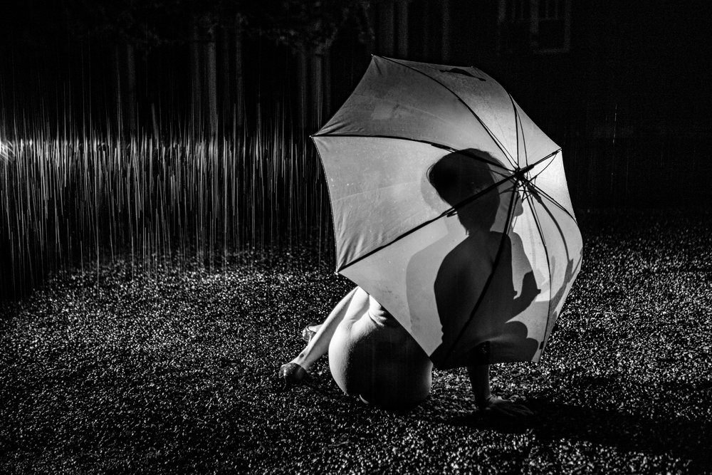 Nude woman sits on wet ground at night holding an umbrella as rain falls, cosplay, nudity, freedom, expression, USA.