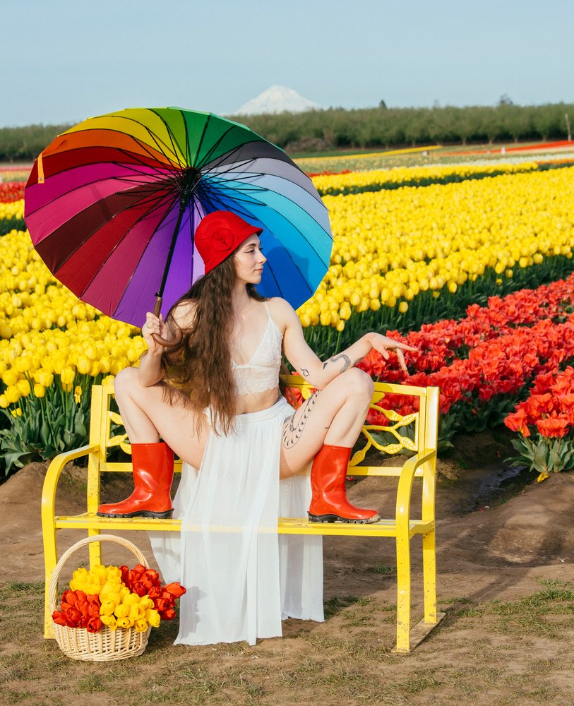 Woman sits on a yellow bench holding a rainbow umbrella amid red and yellow tulip fields with a mountain backdrop, USA.