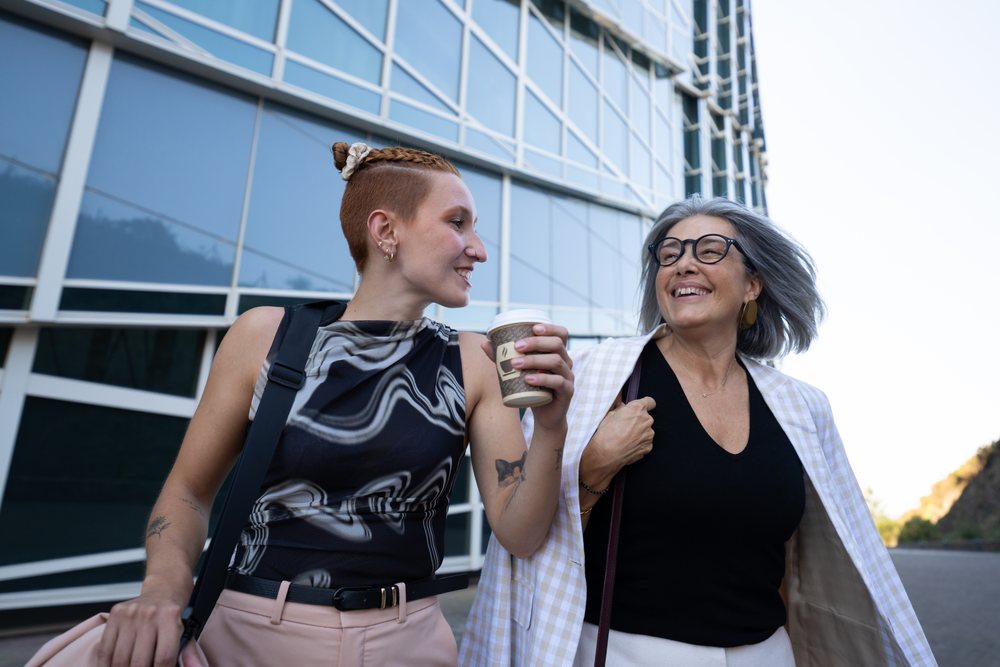 Two businesswomen smiling and talking while walking outside a modern office building, Spain.