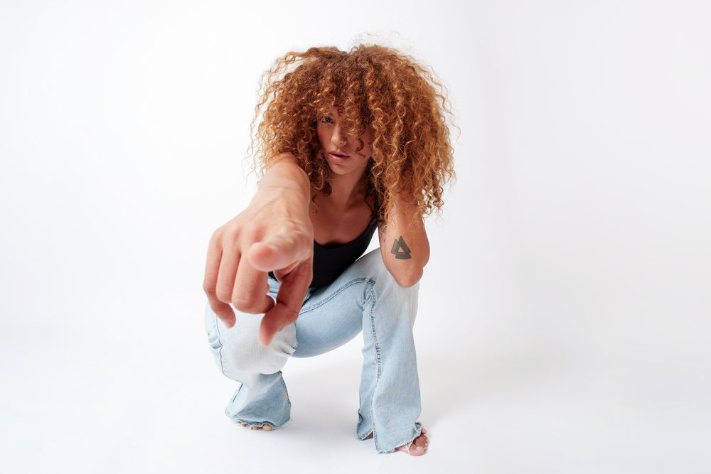 Woman with curly red hair crouching barefoot in a studio against a white background, pointing forward and showing a tattoo on her left upper arm, Malaga, Spain.