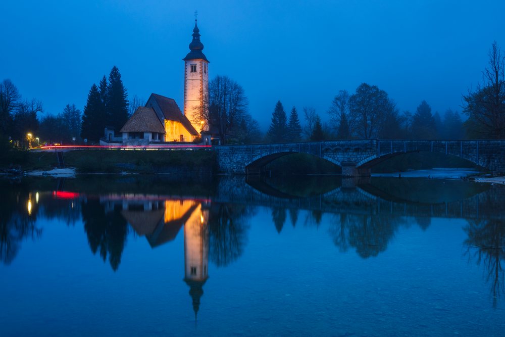 The Church of St John the Baptist is illuminated at twilight, reflecting in the river next to a stone arch bridge, Landscape, Bohinj, Slovenia.
