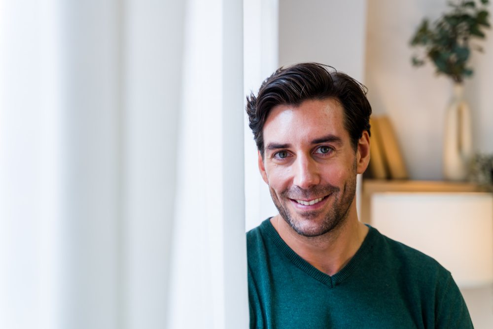Man with brown hair smiling indoors beside a white curtain in a modern Spanish home interior with a lamp, plant, and books visible, Spain.
