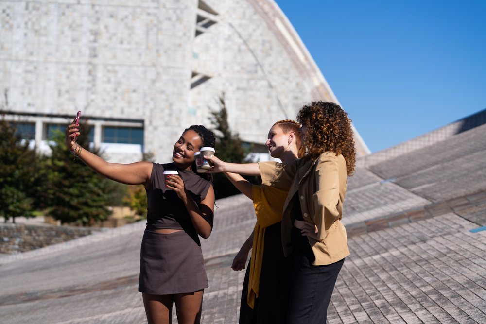 Three women take a selfie outdoors holding coffee cups by a modern building on a sunny day, Spain.
