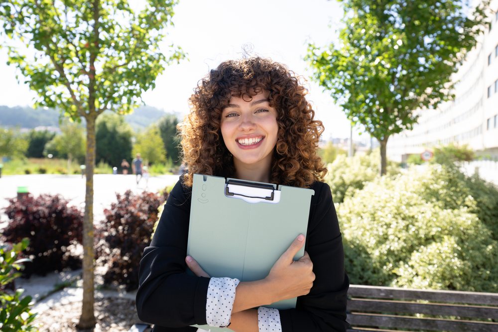 Smiling woman with curly hair holding a document folder while sitting on a bench in a sunlit outdoor park, Spain.