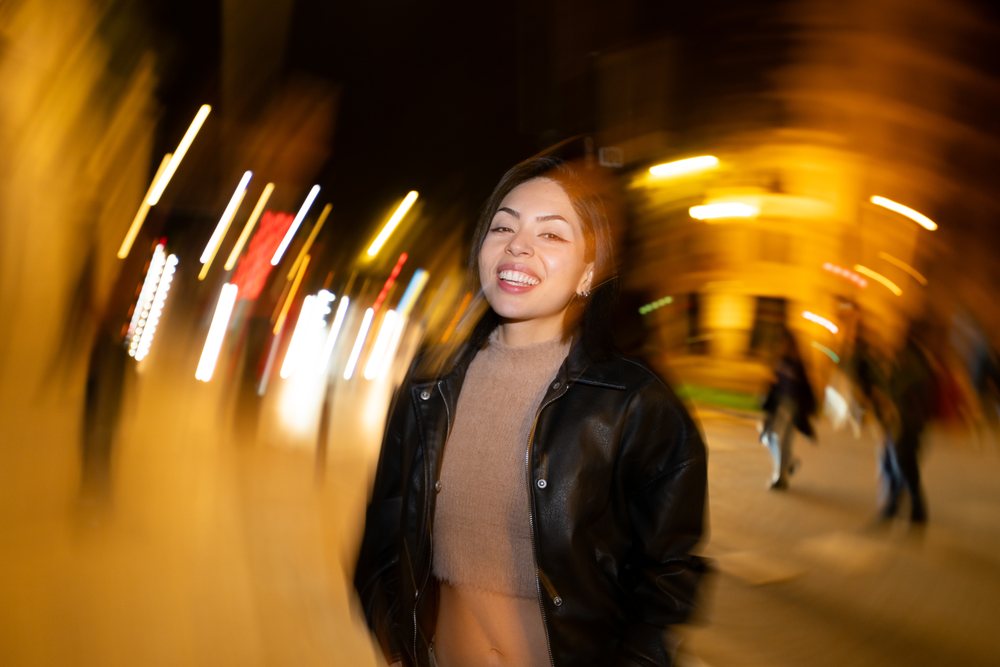 Woman wearing a black leather jacket and brown sweater smiles on a lively city street at night with people and lights behind her, Madrid, Spain.