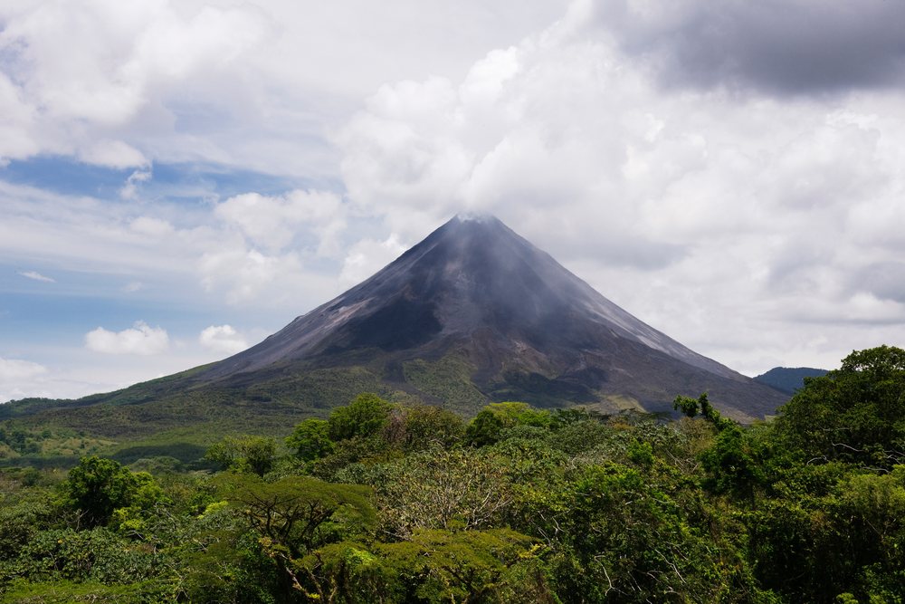 Arenal Volcano rises above lush tropical forest under a cloudy sky in Costa Rica.