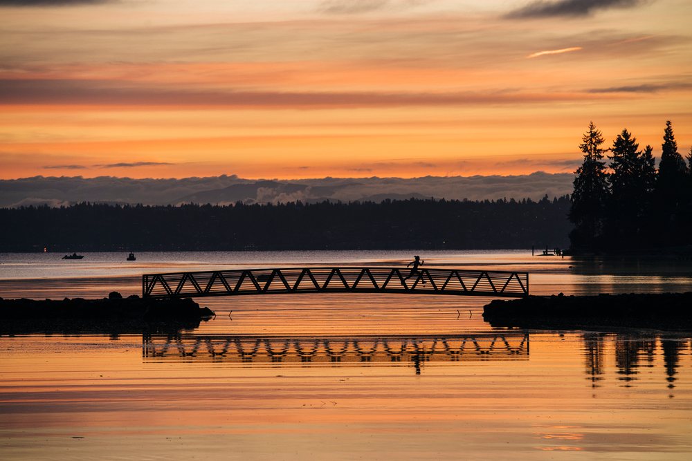 Silhouette of a runner crossing a bridge over calm water reflecting a colorful sunset, with trees and boats visible, Bainbridge Island, WA, USA.