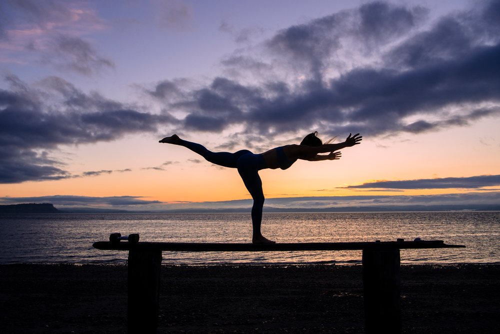 Woman performing a yoga balance pose in silhouette on an elevated wooden plank at sunset on a beach with clouds over water, WA, USA.