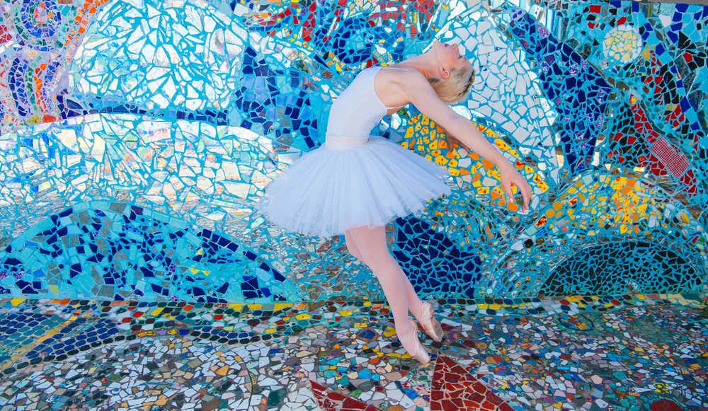 Ballet dancer wearing white tutu and pink tights poses gracefully before a colorful mosaic wall and floor outdoors in California, USA.