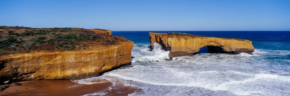 London Bridge rock formation with ocean waves crashing along the coastline, Victoria Australia under a blue sky, Port Campbell National Park.