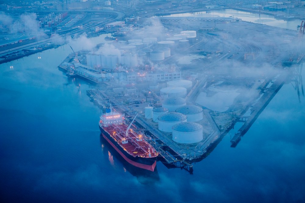Oil tanker ship docked at a fuel storage terminal with multiple oil storage tanks and industrial facilities at the Port of Long Beach, USA under foggy conditions, CA.