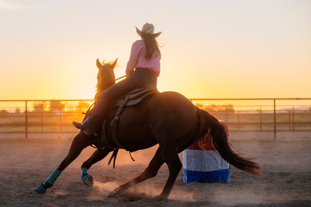 Horse rider wearing a western hat makes a sharp turn around a barrel in a fenced outdoor arena at sunset, Southwest USA.