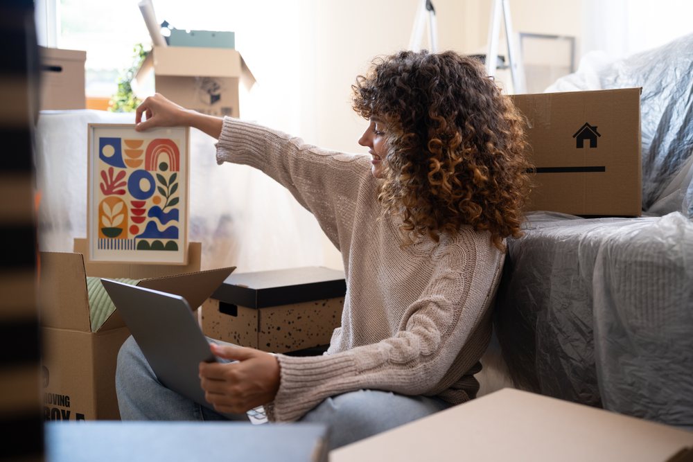 Woman sitting indoors in Spain holds framed abstract art and a laptop, moving.
