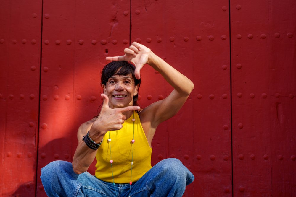 Woman wearing a yellow tank top and jeans smiles while framing her face with her hands in front of a red door, retro, vintage, Málaga, Spain.