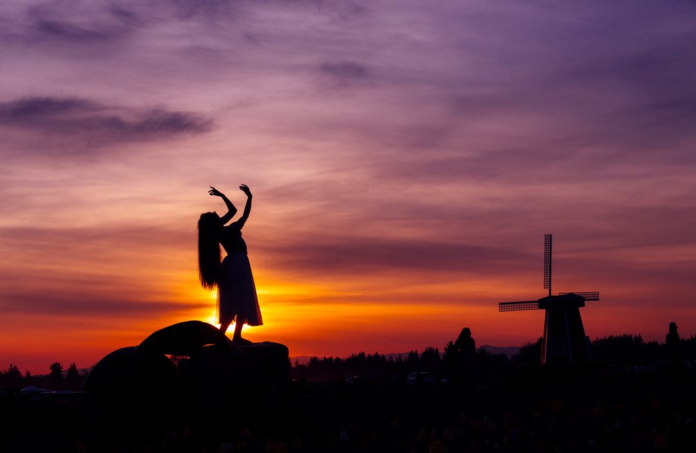 Silhouette of a woman dancing near a windmill at sunset with a colorful sky and nature backdrop, USA.