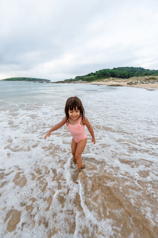 Child in a swimsuit plays in shallow ocean water with waves on a sandy beach backed by green hills under a cloudy sky, kids, Spain.