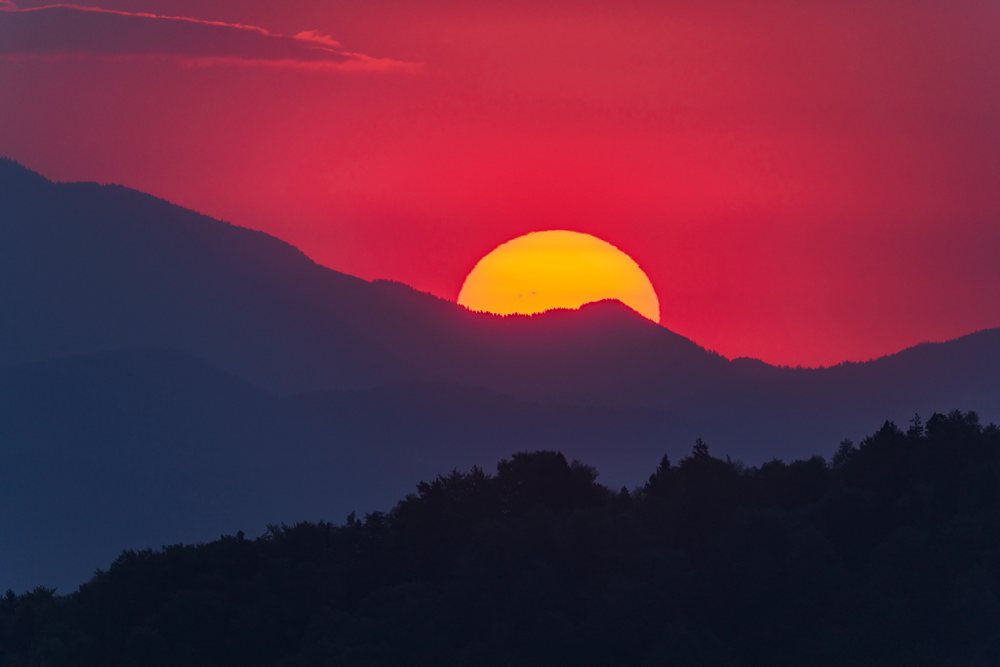 Sun rises behind silhouetted mountains with a vibrant red and orange sky, landscape, cityscape, Skofja Loka, Slovenia.