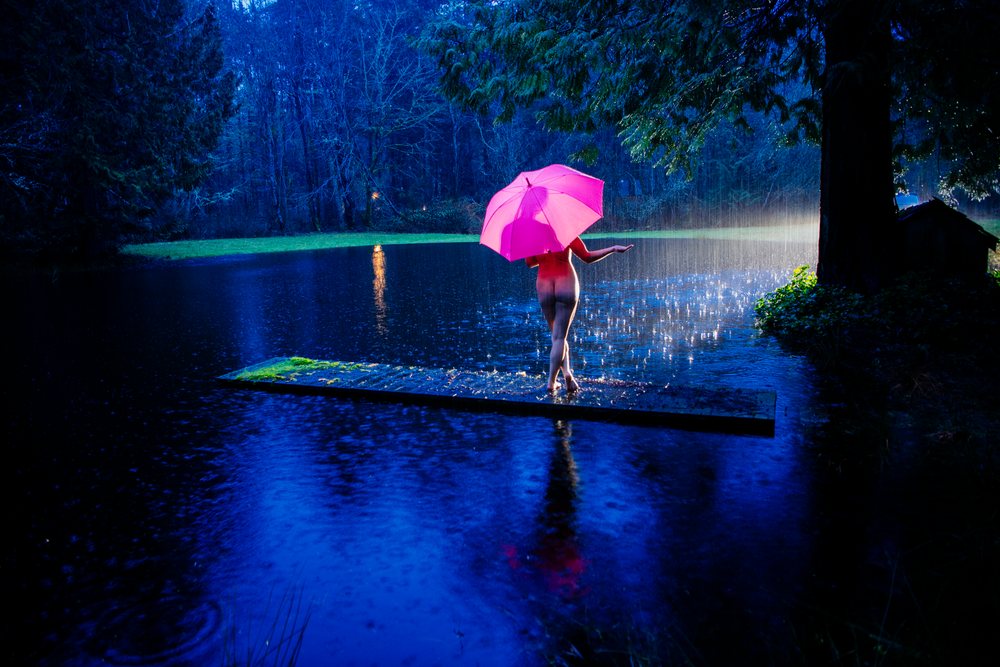 Naked woman holding a pink umbrella stands barefoot on a wooden dock over a pond at night in the USA during rain, cosplay, nudity, freedom, expression.