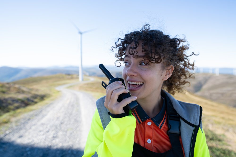 Woman holding a walkie talkie near wind turbines on a sunny day in rural, sustainability, renewable energy, Galicia, Spain.