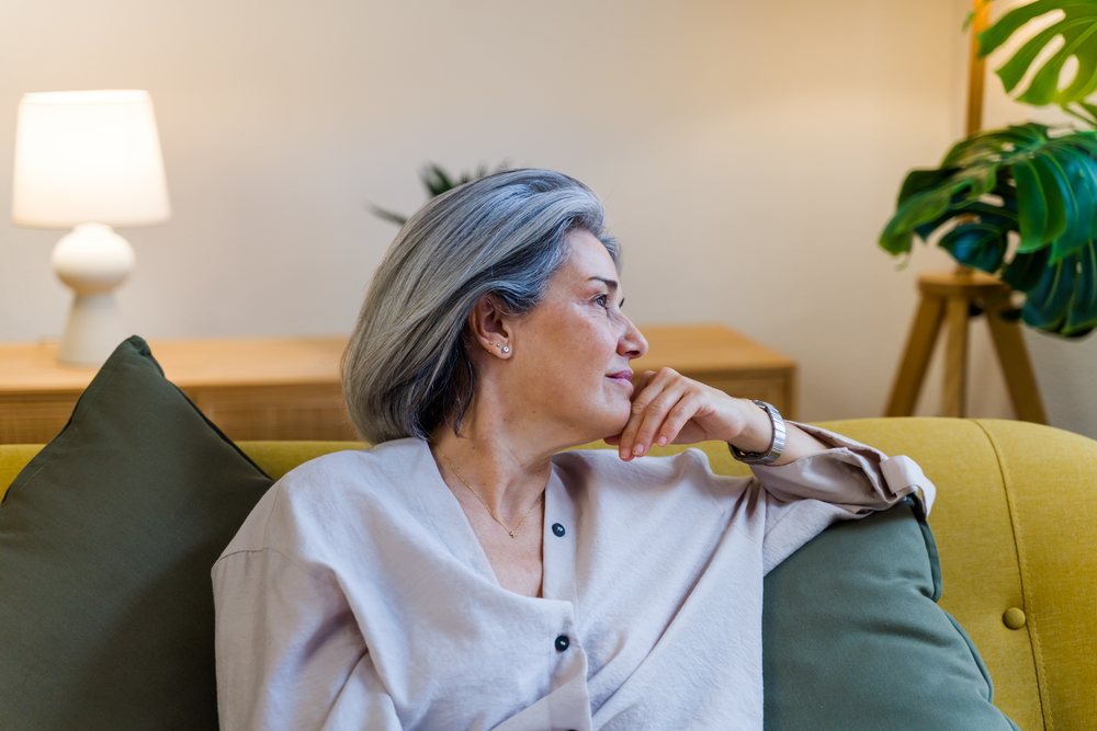 Mature woman with gray hair seated thoughtfully on a yellow sofa with green pillows in a cozy living room with indoor plants and a table lamp, Spain.