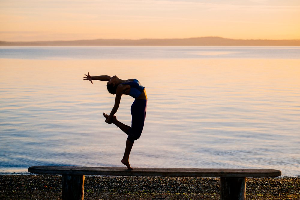 Woman performs a balanced acrobatic yoga pose on a wooden bench by the water at sunset, USA.