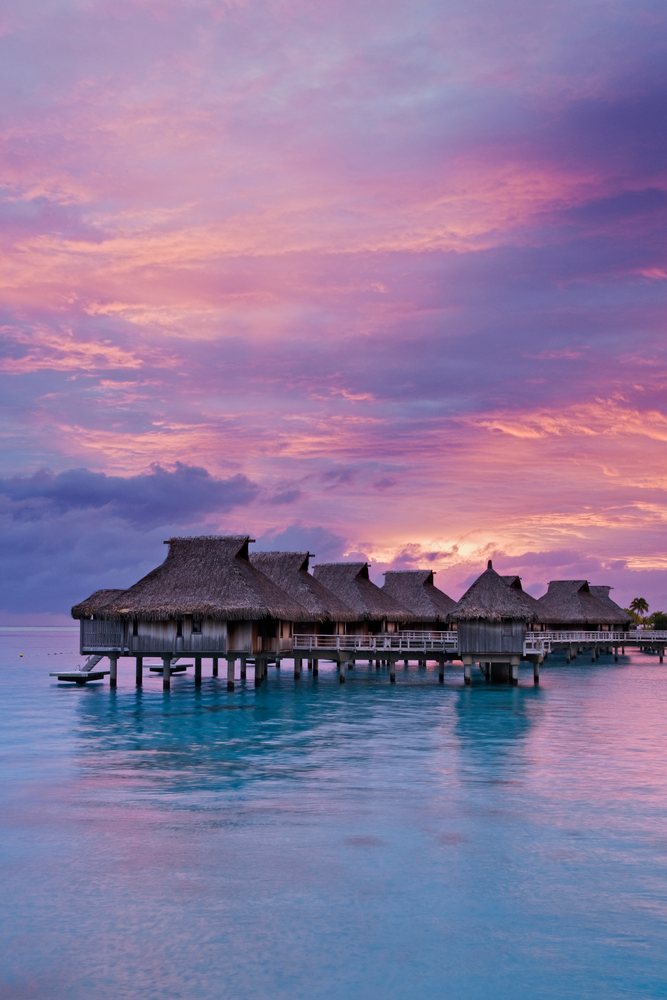 Overwater bungalows with thatched roofs at Bora Nui Resort during a vivid purple sunset over calm ocean waters, Tahiti, Society Islands, Polynesia.
