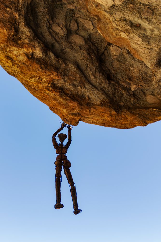 Human-like wire and stone sculpture hanging by hands from a large rock boulder against a clear blue sky, landscape, wildlife, Kunene Region, Namibia.