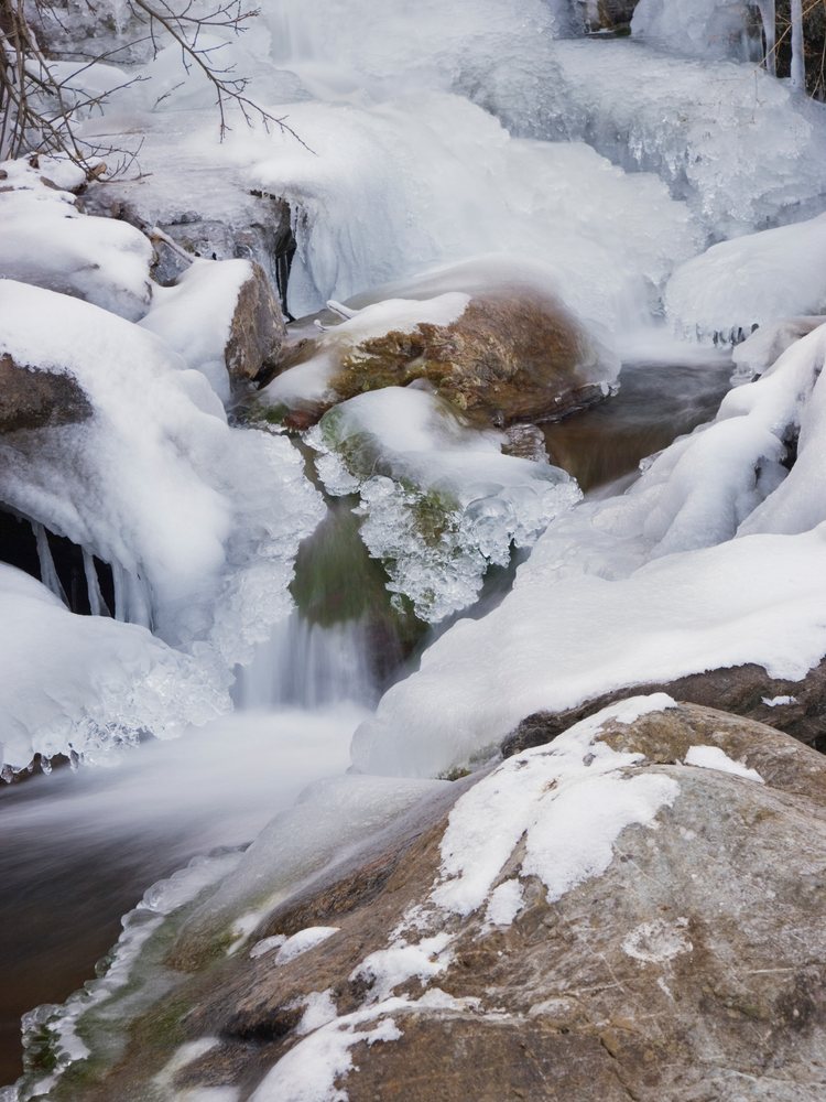 Frozen winter stream flows over snow-covered rocks with ice and icicles visible in a natural outdoor setting, Japan.