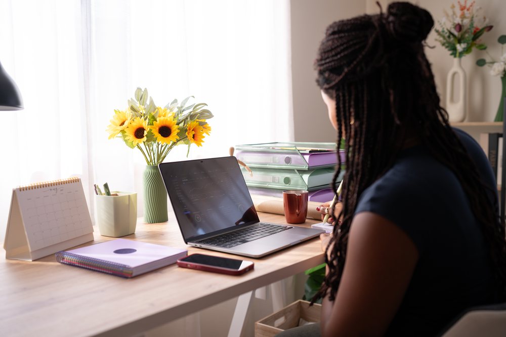 A woman with braided hair works at a home office desk with a laptop, notebooks, and sunflowers, remote work, student, Spain.