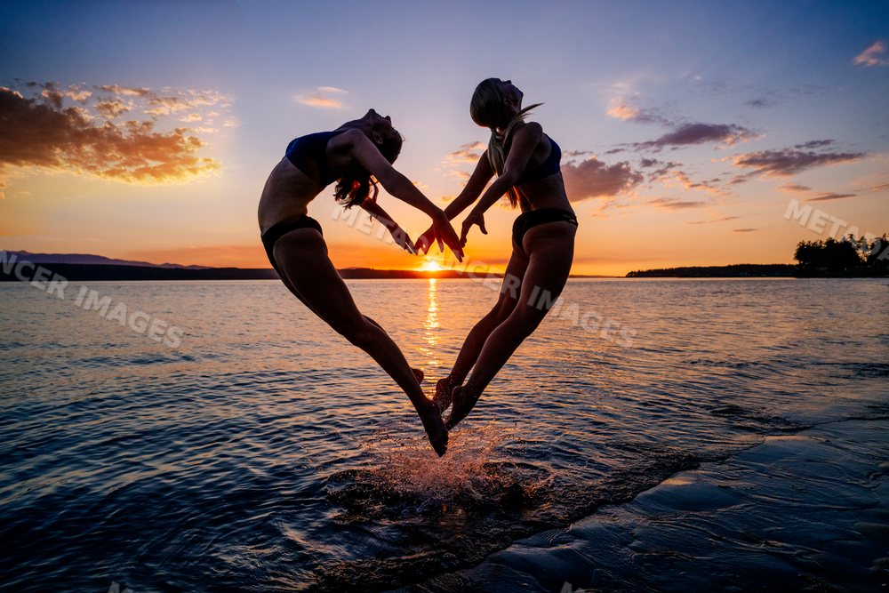 Two white women jump and practice partner acrobatics yoga together