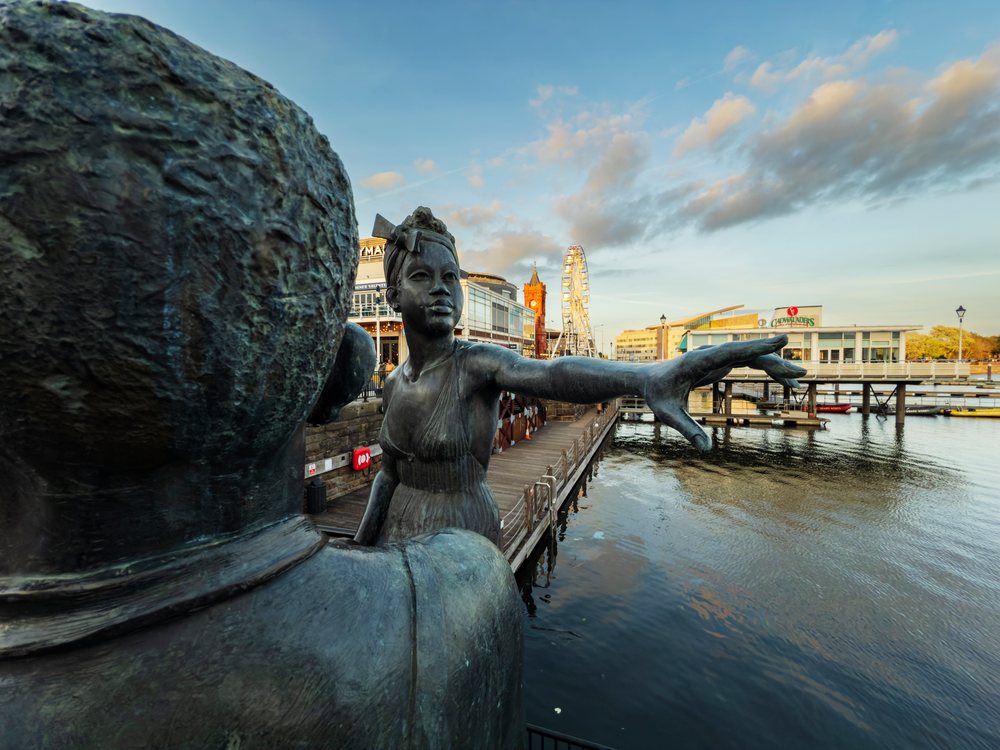 Bronze People Like Us Sculpture with female figure extending hand at Mermaid Quay waterfront in Cardiff under a partly cloudy sky, Landscape, Glamorgan, Wales.