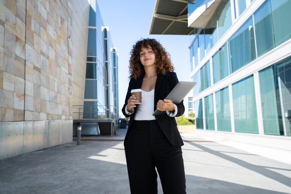 Businesswoman holding a coffee cup and a laptop, standing outside modern office buildings on a sunny day, traveling executive, Spain.