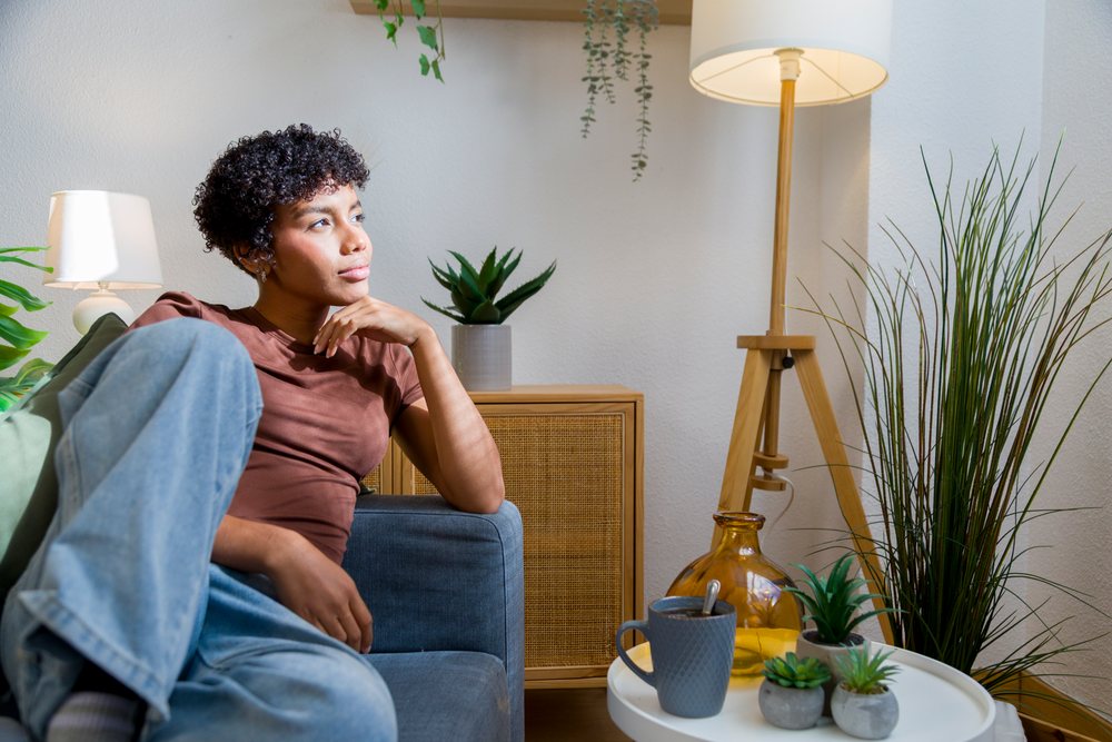 Young woman seated thoughtfully on a gray sofa in a modern living room with indoor plants and a coffee cup on a round white table.