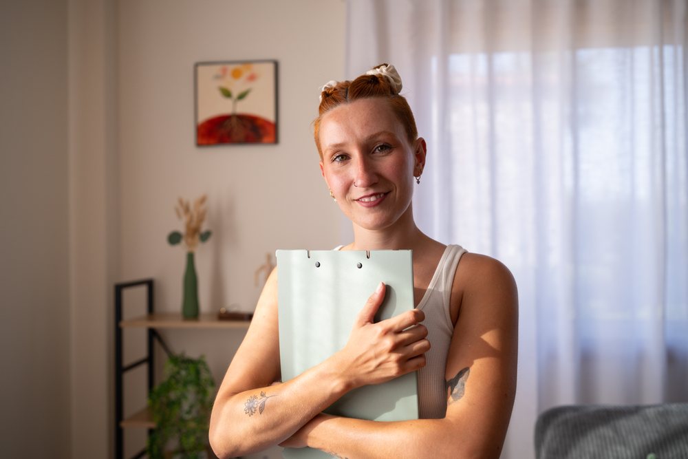 A smiling woman with red hair hugs a folder in a bright home office with plants and wall art, moving, Spain.