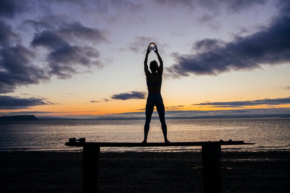 Silhouette of a woman holding a glass ball above her head on a wooden structure at sunset on a beach, WA, USA.