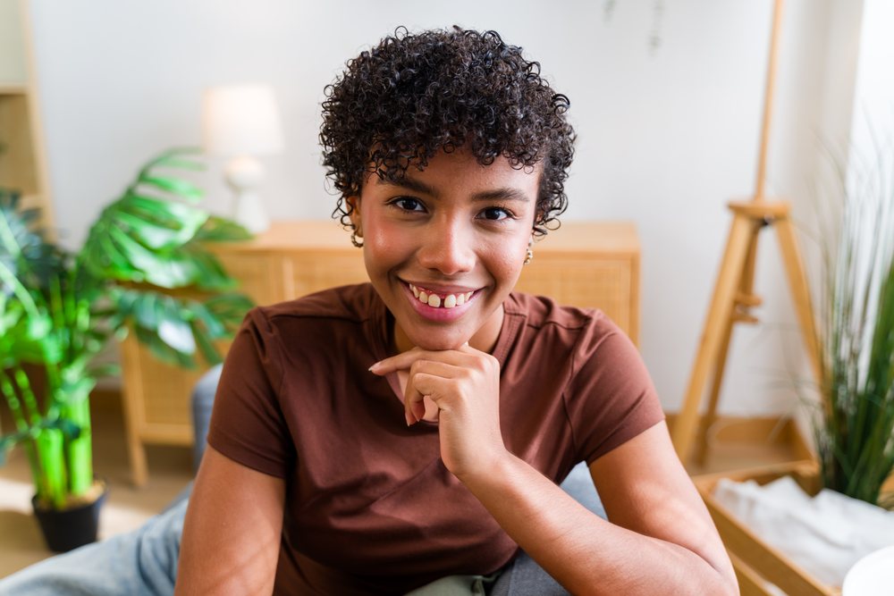 Smiling woman with curly hair wearing a brown shirt sitting in a decorated living room with houseplants and wooden furniture, Spain.